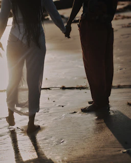 A candid shot of a couple holding hands, walking barefoot along a shoreline with charcoal shadows.