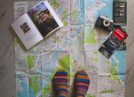A map of Copenhagen is spread out on a wooden floor. An open book with images and text lies on the left side of the map. A vintage camera is positioned on the top right corner of the map, and a travel guidebook titled 'Welcome to Copenhagen' is placed slightly below the camera. A person wearing colorful striped socks is standing on the bottom of the map, adding a personal touch to the scene.