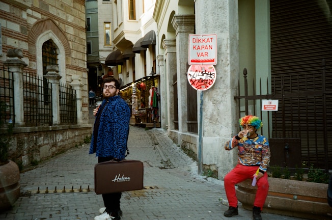 A person wearing a blue patterned coat holds a suitcase labeled 'Helena' while standing in a city street next to a stone building. Another person sitting nearby is dressed in a colorful outfit with a rainbow wig, drinking from a cup. A Turkish warning sign attached to a pole reads 'DIKKAT KAPAN VAR,' and there are graffiti markings underneath.