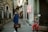 A person wearing a blue patterned coat holds a suitcase labeled 'Helena' while standing in a city street next to a stone building. Another person sitting nearby is dressed in a colorful outfit with a rainbow wig, drinking from a cup. A Turkish warning sign attached to a pole reads 'DIKKAT KAPAN VAR,' and there are graffiti markings underneath.