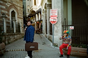 A person wearing a blue patterned coat holds a suitcase labeled 'Helena' while standing in a city street next to a stone building. Another person sitting nearby is dressed in a colorful outfit with a rainbow wig, drinking from a cup. A Turkish warning sign attached to a pole reads 'DIKKAT KAPAN VAR,' and there are graffiti markings underneath.