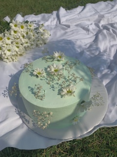 A pastel green sage-themed cake decorated with delicate white flowers on a minimalist wooden table.
