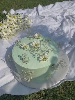 A pastel green sage-themed cake decorated with delicate white flowers on a minimalist wooden table.