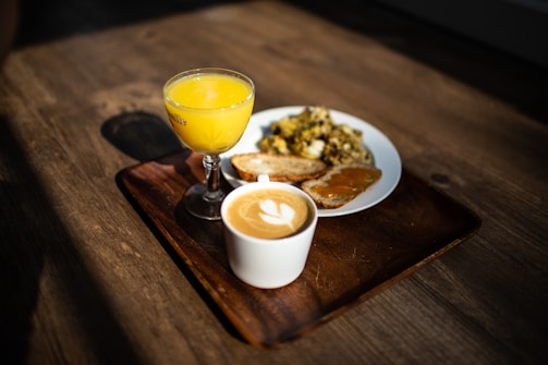 A breakfast setting with a cup of cappuccino featuring latte art, a small glass of orange juice, and a plate containing scrambled eggs with herbs, toast, and toast with what appears to be apricot jam, all placed on a wooden serving tray on a wooden table.