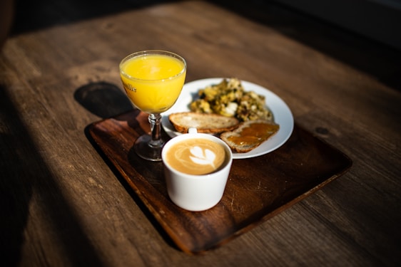 A breakfast setting with a cup of cappuccino featuring latte art, a small glass of orange juice, and a plate containing scrambled eggs with herbs, toast, and toast with what appears to be apricot jam, all placed on a wooden serving tray on a wooden table.
