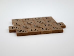 Wooden Scrabble tiles are arranged to form the phrase 'TRUE LOVE NEEDS WORK' on a light surface.
