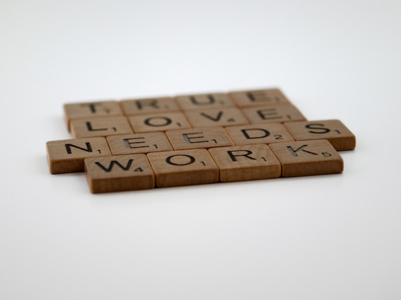 Wooden Scrabble tiles are arranged to form the phrase 'TRUE LOVE NEEDS WORK' on a light surface.