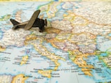 A small, vintage toy airplane rests on a detailed map of Europe, with visible countries and cities including Spain, France, Italy, and Greece. The map is rich in colors, with borders and text indicating geographical distinctions.