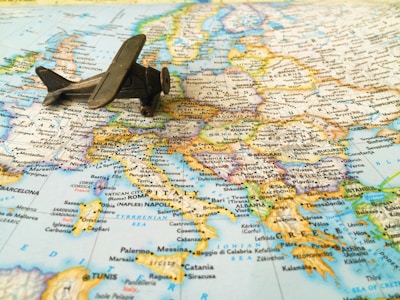 A small, vintage toy airplane rests on a detailed map of Europe, with visible countries and cities including Spain, France, Italy, and Greece. The map is rich in colors, with borders and text indicating geographical distinctions.