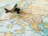 A small, vintage toy airplane rests on a detailed map of Europe, with visible countries and cities including Spain, France, Italy, and Greece. The map is rich in colors, with borders and text indicating geographical distinctions.
