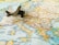 A small, vintage toy airplane rests on a detailed map of Europe, with visible countries and cities including Spain, France, Italy, and Greece. The map is rich in colors, with borders and text indicating geographical distinctions.