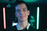 A young person stands in front of a dimly lit background featuring neon lights in red and blue. The focus is on the individual, who is wearing a dark shirt with a white collar. The setting suggests a modern and tech-inspired environment with a moody ambiance.