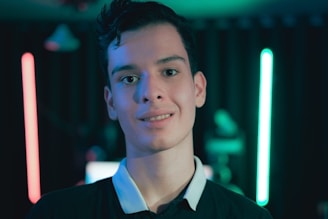 A young person stands in front of a dimly lit background featuring neon lights in red and blue. The focus is on the individual, who is wearing a dark shirt with a white collar. The setting suggests a modern and tech-inspired environment with a moody ambiance.