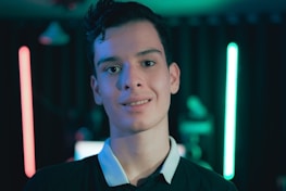 A young person stands in front of a dimly lit background featuring neon lights in red and blue. The focus is on the individual, who is wearing a dark shirt with a white collar. The setting suggests a modern and tech-inspired environment with a moody ambiance.