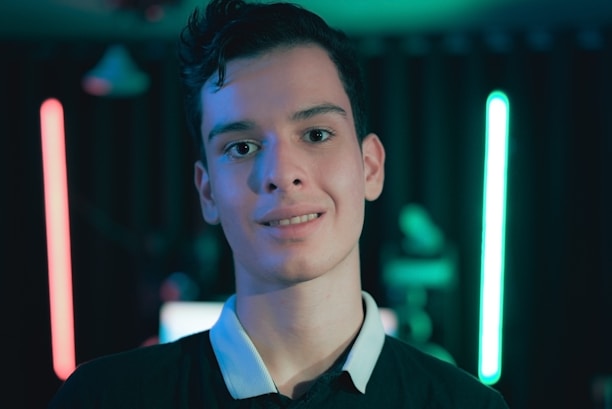 A young person stands in front of a dimly lit background featuring neon lights in red and blue. The focus is on the individual, who is wearing a dark shirt with a white collar. The setting suggests a modern and tech-inspired environment with a moody ambiance.