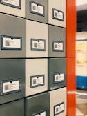 A series of square lockers with QR codes and labels featuring names in Cyrillic script. The lockers are arranged in a grid pattern with a mix of gray and white colors. A bold red vertical divider is visible, and a blurred background suggests a public space, possibly a train station.