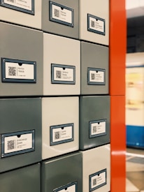 A series of square lockers with QR codes and labels featuring names in Cyrillic script. The lockers are arranged in a grid pattern with a mix of gray and white colors. A bold red vertical divider is visible, and a blurred background suggests a public space, possibly a train station.