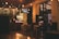 A cozy café interior with wooden furniture and warm lighting. Decorative light bulbs hang from the ceiling, and a menu is displayed above the counter. A neon sign shows a price in UAH. Outside, there are visible reflections and a glimpse of a city street through large windows.