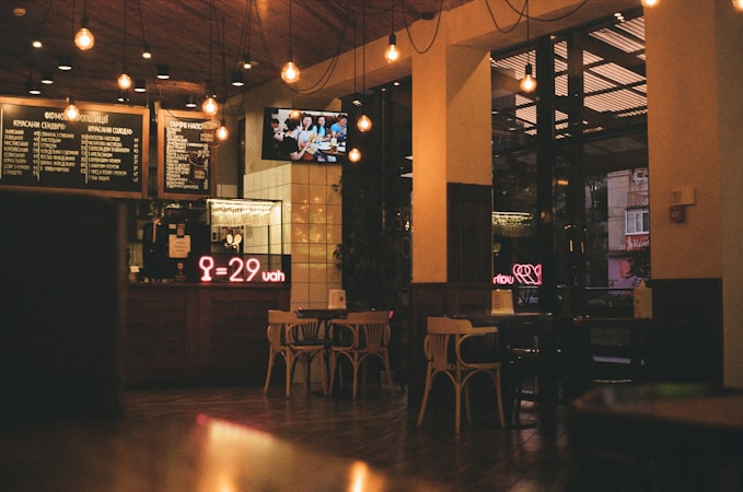 A cozy caf&eacute; interior with wooden furniture and warm lighting. Decorative light bulbs hang from the ceiling, and a menu is displayed above the counter. A neon sign shows a price in UAH. Outside, there are visible reflections and a glimpse of a city street through large windows.