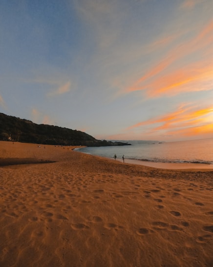 A serene beach at sunset with golden sands and the sky painted in hues of orange and blue. The coastline curves gently into the distance, with a couple of silhouettes walking along the shore. The sea is calm, and the horizon blends seamlessly into the sky.
