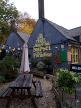 A quaint outdoor seating area features a wooden picnic table with an umbrella. The background shows a building with a dark facade displaying a phrase in large yellow letters. There are plants and shrubs arranged around the seating area, and colorful bunting hangs across the space. The atmosphere is cozy and inviting, suggesting a setting for relaxation or social gathering.
