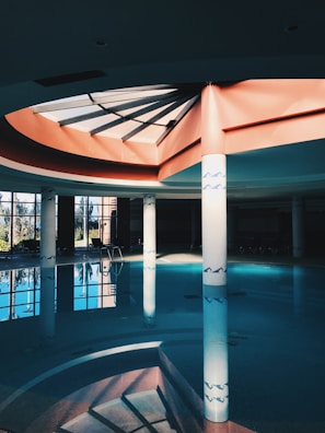 A spacious indoor swimming pool area with large glass windows, allowing natural light to create reflections on the water. The ceiling design features a geometric skylight with beams that casts shadows on the walls. White columns with decorative blue patterns support the structure, and several lounge chairs are visible around the pool area.