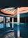 A spacious indoor swimming pool area with large glass windows, allowing natural light to create reflections on the water. The ceiling design features a geometric skylight with beams that casts shadows on the walls. White columns with decorative blue patterns support the structure, and several lounge chairs are visible around the pool area.