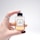 A hand with burgundy painted nails and a silver ring holds a small, square-shaped perfume bottle. The bottle is labeled 'N°3 THE SILENCE PERFUME' and contains a light amber liquid. The background is a soft, neutral gray.