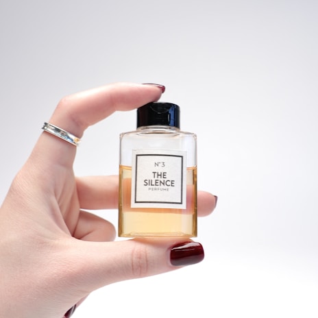 A hand with burgundy painted nails and a silver ring holds a small, square-shaped perfume bottle. The bottle is labeled 'N°3 THE SILENCE PERFUME' and contains a light amber liquid. The background is a soft, neutral gray.