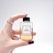 A hand with burgundy painted nails and a silver ring holds a small, square-shaped perfume bottle. The bottle is labeled 'N°3 THE SILENCE PERFUME' and contains a light amber liquid. The background is a soft, neutral gray.