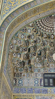 Intricate Islamic architectural tile work featuring elaborate geometric patterns and Arabic calligraphy. The mosaic is primarily composed of blues, golds, and whites, creating a rich tapestry of colors. The arch under which this design is placed is finely adorned, demonstrating traditional artistry.