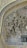 Intricate Islamic architectural tile work featuring elaborate geometric patterns and Arabic calligraphy. The mosaic is primarily composed of blues, golds, and whites, creating a rich tapestry of colors. The arch under which this design is placed is finely adorned, demonstrating traditional artistry.