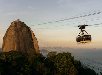 A cable car is traveling towards a large, iconic granite peak surrounded by lush greenery, with a vast ocean and small islands visible in the distance under a clear sky.