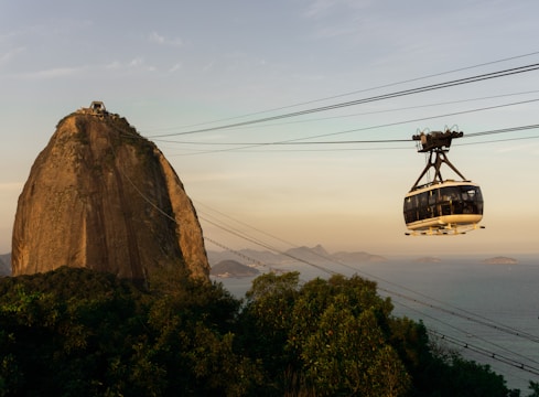 A cable car is traveling towards a large, iconic granite peak surrounded by lush greenery, with a vast ocean and small islands visible in the distance under a clear sky.