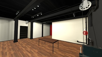 A modern, minimalist interior with wooden flooring and black metal structural beams. The space features a simple table, a dark cabinet, and a wall with a large white surface. Industrial elements are prominent, including exposed pipes and a large, red valve.