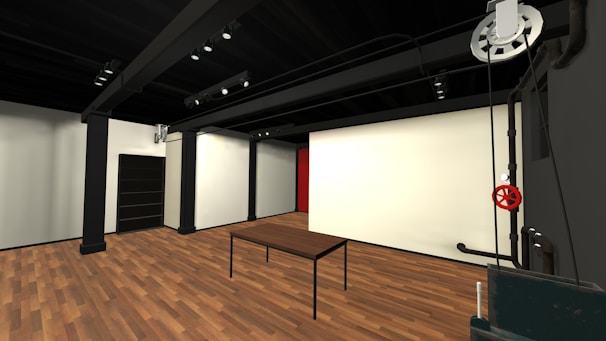 A modern, minimalist interior with wooden flooring and black metal structural beams. The space features a simple table, a dark cabinet, and a wall with a large white surface. Industrial elements are prominent, including exposed pipes and a large, red valve.