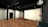 A modern, minimalist interior with wooden flooring and black metal structural beams. The space features a simple table, a dark cabinet, and a wall with a large white surface. Industrial elements are prominent, including exposed pipes and a large, red valve.