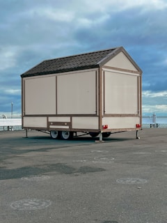 A small beige-colored mobile structure with a gabled roof is placed on a paved area near a waterfront. It is mounted on a two-axle trailer. The background features a cloudy sky and a glimpse of a body of water, along with some railings and a few distant lampposts.