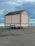 A small beige-colored mobile structure with a gabled roof is placed on a paved area near a waterfront. It is mounted on a two-axle trailer. The background features a cloudy sky and a glimpse of a body of water, along with some railings and a few distant lampposts.