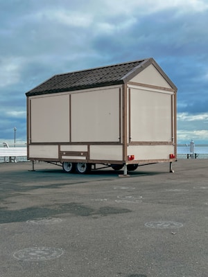 A small beige-colored mobile structure with a gabled roof is placed on a paved area near a waterfront. It is mounted on a two-axle trailer. The background features a cloudy sky and a glimpse of a body of water, along with some railings and a few distant lampposts.