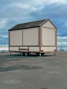 A small beige-colored mobile structure with a gabled roof is placed on a paved area near a waterfront. It is mounted on a two-axle trailer. The background features a cloudy sky and a glimpse of a body of water, along with some railings and a few distant lampposts.