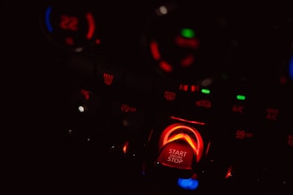 Close-up of a car's dashboard with illuminated control buttons. A prominent start/stop engine button glows in red along with various symbols and temperature controls. Dim lighting emphasizes the colorful display and creates a high contrast.