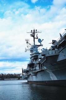 A large naval battleship, possibly an aircraft carrier, is docked on a body of water. The ship is equipped with various antennas and radar systems on its deck and superstructure. The sky is partly cloudy with patches of blue, and there are trees and a few buildings visible in the distance along the shoreline.