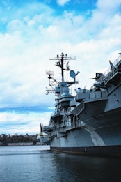 A large naval battleship, possibly an aircraft carrier, is docked on a body of water. The ship is equipped with various antennas and radar systems on its deck and superstructure. The sky is partly cloudy with patches of blue, and there are trees and a few buildings visible in the distance along the shoreline.