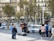 A street musician plays an acoustic guitar in a plaza, surrounded by several children who are watching him perform. The area is lined with trees and benches, with additional people sitting in the background. Buildings with balconies form the backdrop, creating an urban environment.