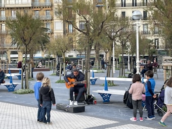 A street musician plays an acoustic guitar in a plaza, surrounded by several children who are watching him perform. The area is lined with trees and benches, with additional people sitting in the background. Buildings with balconies form the backdrop, creating an urban environment.