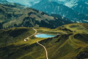 A serene mountain landscape with rolling green hills and a small lake in the center. Winding paths lead towards the lake, surrounded by vibrant greenery and clusters of pine trees. The background features expansive mountain ranges, creating a sense of depth and grandeur.