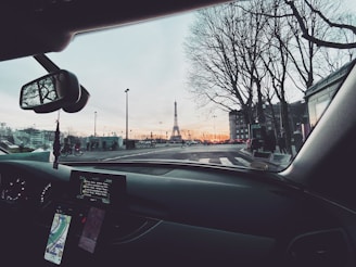 A view from inside a car showing the dashboard with navigation screens and a rearview mirror. Outside the car, the Eiffel Tower is visible in the distance, surrounded by tall trees and streetlights. The sky is a mix of soft colors from the sunset.