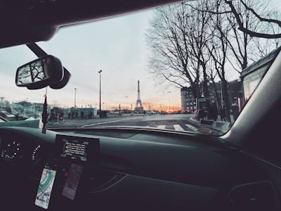 A view from inside a car showing the dashboard with navigation screens and a rearview mirror. Outside the car, the Eiffel Tower is visible in the distance, surrounded by tall trees and streetlights. The sky is a mix of soft colors from the sunset.