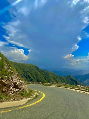 A winding mountain road curves into the distance, bordered by rocky terrain and lush green hills. Above, a vast sky showcases dramatic clouds with soft, bright tones of white and gray. The landscape is expansive, providing a sense of depth and natural grandeur.